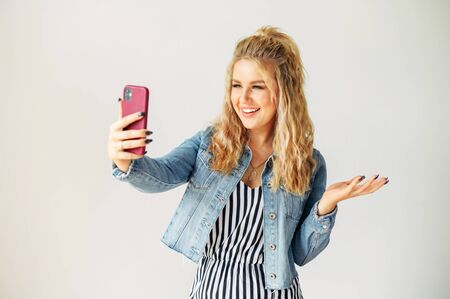 Attractive young blonde woman using fancy smartphone for video call. Woman is striped dress stands and watches at selfie cam with a smile, white wall on backgroundの写真素材