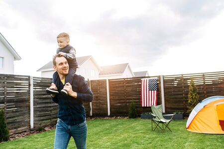 Camping in the backyard. A dad and son set up a tent on a lawn and they are spending time funny together. A boy sits on the shoulders of dad and laughsの写真素材