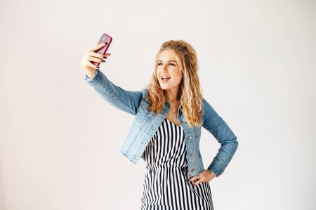 Selfie time. Cheerful young blonde woman is taking selfie on a trendy smartphone with a white wall on backgroundの写真素材