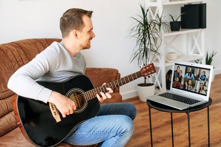 Online musical performance. A young guy sits on the sofa at home and plays acoustic guitar in front of online audience on the laptop screen. Video concert online, video chatの写真素材