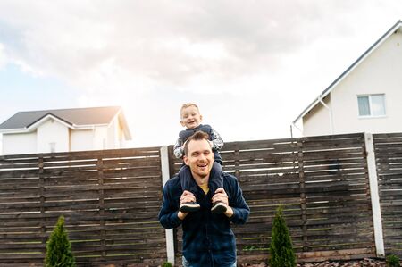 Father and son are happy to spend time together outdoors. Dad is carrying his little son on the shoulders, they laughの写真素材