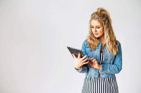 Young beautiful woman with a curly blonde hair is using tablet computer while standing with a white wall on backgroundの写真素材