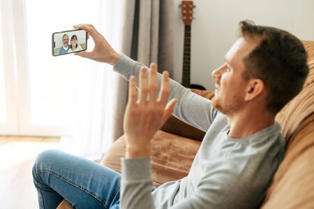 A guy is talking online with a parents, he sits on the sofa and using smartphone for online communication. Virtual meeting with a familyの写真素材