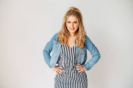 Pure beauty. Beautiful young blonde woman in striped dress and in jeans jacket looks at camera with a smile, she stands in pose hands on hips. Isolated on white backgroundの写真素材
