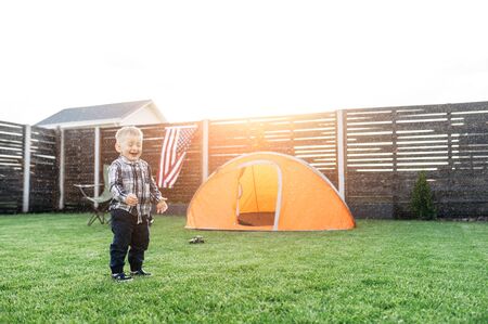 Cute baby boy stands on a lawn under rain drops and laughs. Camping tent on the background. Home leisure in the backyardの写真素材