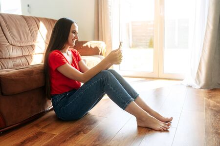 Young woman talks to mom by video link, she is sitting at cozy living room and using smartphone for connectの写真素材