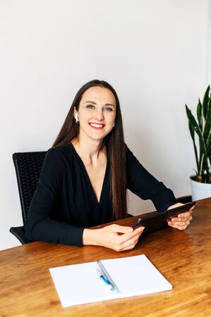 A young beautiful woman is using digital tablet while sitting at the table in office. She is holding tablet in hands, looks at camera and smilesの写真素材