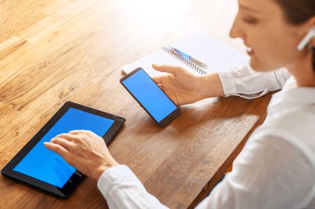 Back view of a young woman is white smart casual shirt sits with a tablet and smartphone in hands at the table indoors. Copy space for inserting text on a blank screens of gadgetsの写真素材