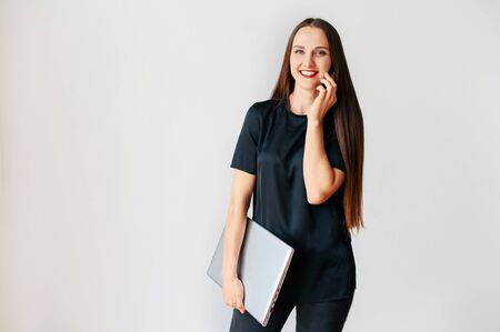 Young cheerful attractive woman in black wear with a laptop is talking on the phone and smiles indoors. Isolated on white backgroundの写真素材