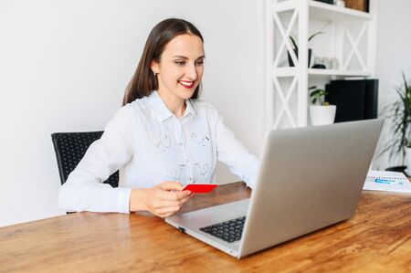 A woman using laptop for online shopping, online banking. She sits at the table with a credit card in hand and looks at the screen with a smileの写真素材