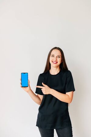 A young cheerful girl in black t-shirt holds a smartphone in hand and points by finger of other hand on the blank screen. Copy space for text. Isolated on white. Gadgets and technology concept.の写真素材