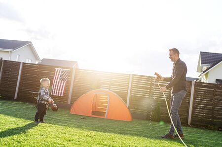 Young father and little cute son spend active time together in the backyard. Dad is watering son from hose, a boy is running away and laughingの写真素材