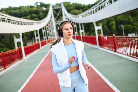 Healthy active lifestyle. Pretty athlete girl in trendy sports wear with a headphones is running on the city bridge in a sunset lightの写真素材