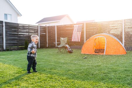 Cute baby boy stands on a lawn under rain drops and laughs. Camping tent on the background. Home leisure in the backyardの写真素材
