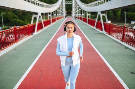 Healthy active lifestyle. Pretty athlete girl in trendy sports wear with a headphones is running on the city bridge in a sunset lightの写真素材