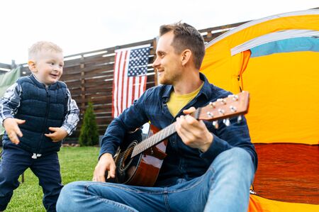 Father and son spend time in the backyard. Daddy plays guitar, toddler son plays nearの写真素材