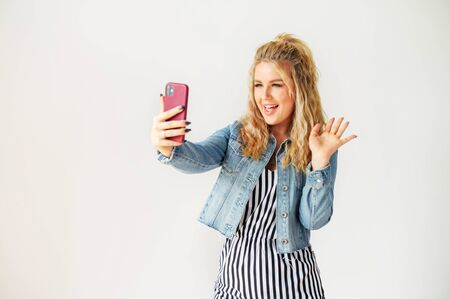 Cheerful young blonde woman using fancy smartphone for video call. Woman is striped dress stands and watches at selfie camera with a smile, white wall on backgroundの写真素材