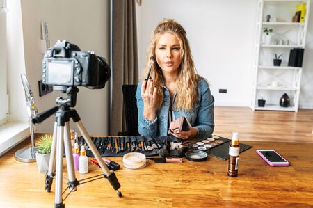 Pretty female beauty blogger is applying powder with a brush and recording makeup video tutorial on a camera at home. Online beauty classesの写真素材