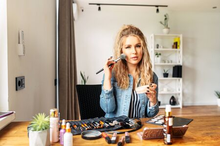 A young well-groomed woman with a smooth skin doing self makeup at home, she sits at the desk and looks at mirror, cosmetics products on the tableの写真素材