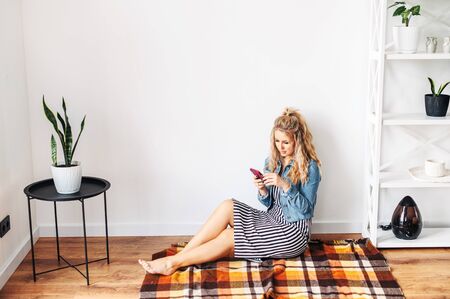 Home leisure with a smartphone. A young girl sits on a cozy plaid in the bright living room and using phone for chatting, web surfing, reading news while staying at homeの写真素材