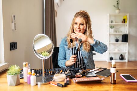 A young and attractive woman sits at a table with cosmetics on it and holds makeup brushes. A professional make-up artist, a make-up teacher.の写真素材
