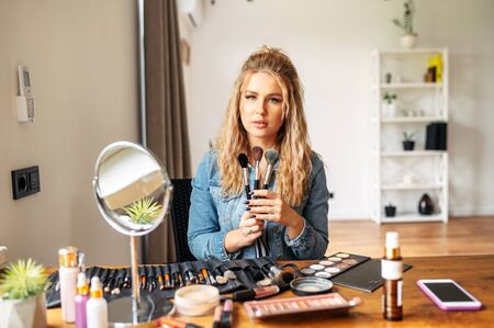 A young and attractive woman sits at a table with cosmetics on it and holds makeup brushes. A professional make-up artist, a make-up teacher.の写真素材