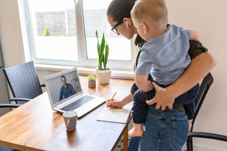 Student mother is watching a lectures, webinars on laptop and writing syllabus with a toddler kid on her arms, male online teacher on the screenの写真素材