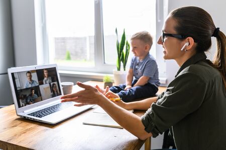 Stay at home. Mother working from home, she talks online with a coworkers via video call on laptop, a cute kid nearの写真素材