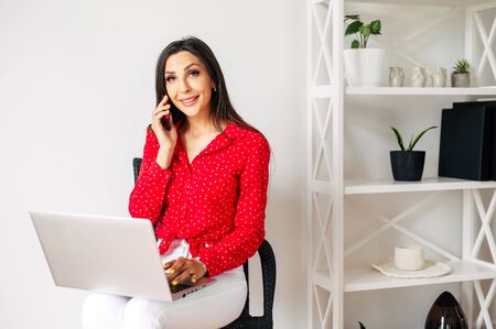 Smiling girl with a phone and laptop on her knees. Remote workplace at home of a business woman. Making business from home, teleworking.の写真素材
