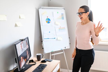 Young girl is laughing and waving hi while a video call with colleagues, cheerful and positive look. Smiling lady with a ponytail and glasses is having a video meeting, standing in home office.の写真素材