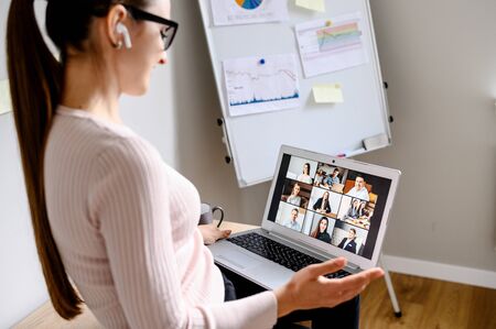 Online video discussion with many people together. Young woman sits in front of laptop with many icons, photo profiles of people on screen. App for video meeting, office with flip chartの写真素材