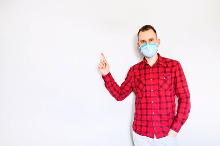 Guy in casual red plaid shirt with surgery protective mask on a face pointing finger at empty space. Isolated on light gray backgroundの写真素材