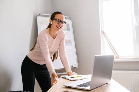 Woman in office has video conversation. Young positive woman in glasses standing in front of laptop and talking with somebody online, flip chart on backgroundの写真素材