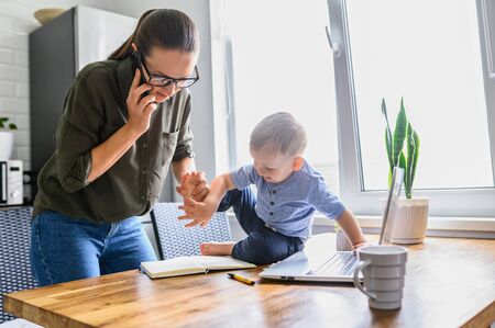 A toddler boy interferes mother with work at home. A young mom in eyeglasses watching on laptop and writes in notebook while kid sits near on a tableの写真素材