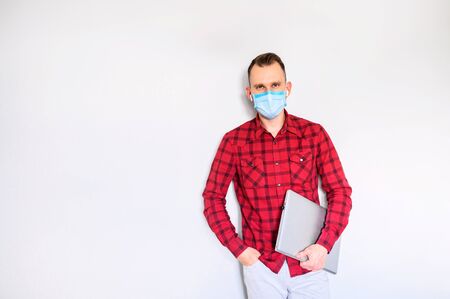 Young guy in casual wear with surgery mask on the face stands with laptop, isolated on gray backgroundの写真素材