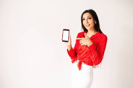 Beautiful young woman with blank smartphone on a white background. Girl in red is pointing finger at blank phone screen. Mockup, copyspace for textの写真素材