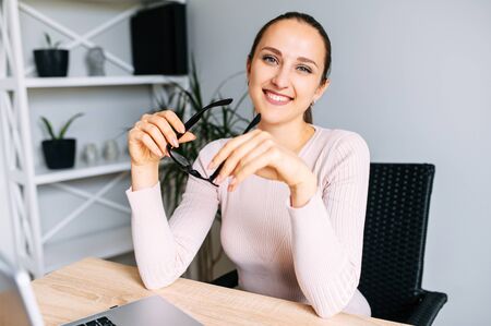 Cheerful young woman in office space. A woman with glasses looks at camera with smile sitting at the desk with a laptopの写真素材