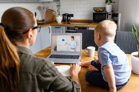 Mom studies online while babysitting with toddler boy. Young mother is watching lecture online and writing syllabus in notebook, her little son sits nearの写真素材