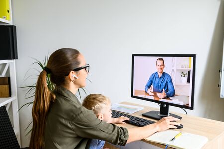 Back view young mother sits with child on laps in home office and talking via video call with male coworker, employee. Mom works at homeの写真素材