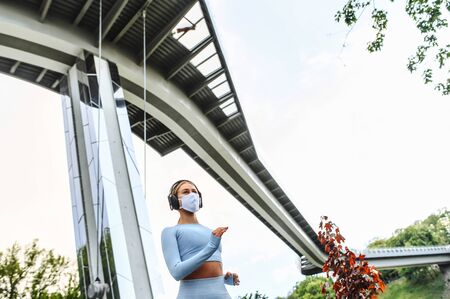 Sports training during quarantine, pandemic. Young woman in protective medical mask running outdoors under city bridgeの写真素材