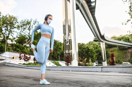 Workout outdoors during quarantine, pandemic. Young woman in protective medical mask doing stretching exercises. Keep your shapeの写真素材