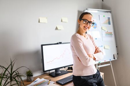 Woman with hands crossed around her waist stands at home office and looks in camera wearing glasses, home workplace and wall with sticky notes on the background.の写真素材