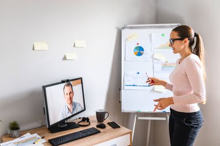 Videocall of young woman with her boss in office, she is standing and presenting some graphs on a flip chart. Casually dressed woman is having a video meeting in office with a colleague, distant work.の写真素材
