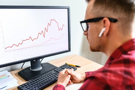 A young serious man looks at the PC screen with graphs on it, he sits at the office desk with hands on it, he is in glasses and headphones, a side view from his backの写真素材