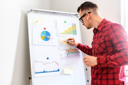 Young concentrated man in glasses with headphone in his ear stands in office, he is making notes on a flipchart with graphs and statistics data on it, a midshotの写真素材