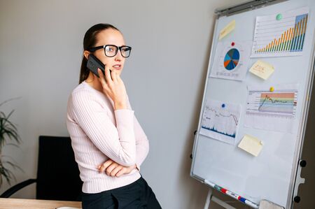 A businesswoman wearing glasses talks on the phone and discusses profit and loss figures for the current period, graphs and diagrams on flip chart . Woman looks away thoughtfulの写真素材