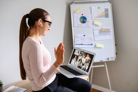 Morning meeting online. A young woman is using app on laptop for connection with colleagues, employees. Video call with many people together. Back viewの写真素材