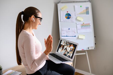 Morning meeting online. A young woman is using app on laptop for connection with colleagues, employees. Video call with many people together. Back view