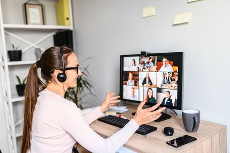 Online meeting, remote work. Young business woman with glasses using app on PC for video communication with many people together, she has headset for talking online with employees. Back viewの写真素材