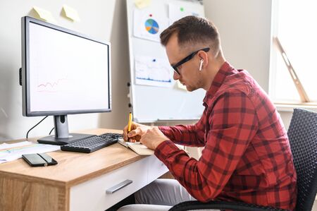 A concentrated guy is making notes into his notebook while sitting at the office desk in front of the computer, he is wearing glasses and headphonesの写真素材